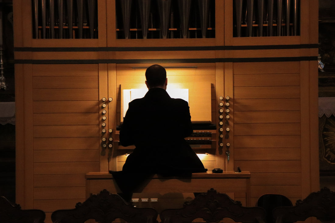 Organist seated at a warmly lit pipe organ console beneath the pipe case, seen from behind