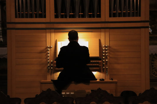 Organist seated at a warmly lit pipe organ console beneath the pipe case, seen from behind