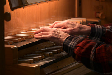 An older organist's weathered hands in a plaid flannel sleeve resting on the wooden manuals of a pipe organ in warm, low light