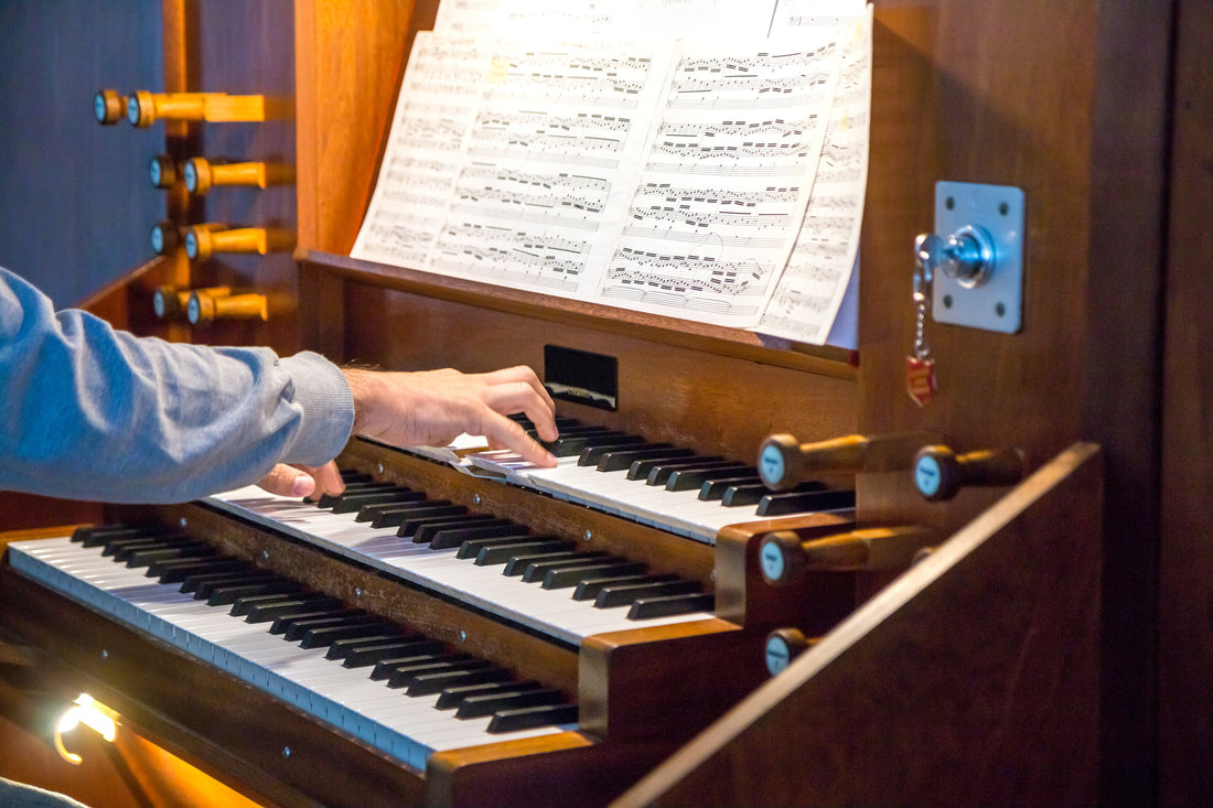 An organist's hand on the upper manual of a warm wooden pipe organ console, with drawknobs at the side and open sheet music on the music rack
