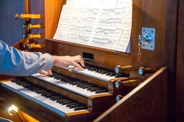 An organist's hand on the upper manual of a warm wooden pipe organ console, with drawknobs at the side and open sheet music on the music rack