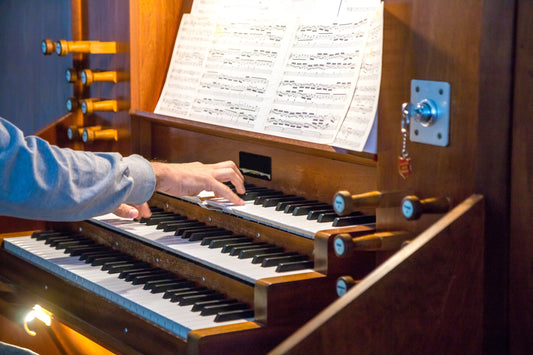 An organist's hand on the upper manual of a warm wooden pipe organ console, with drawknobs at the side and open sheet music on the music rack