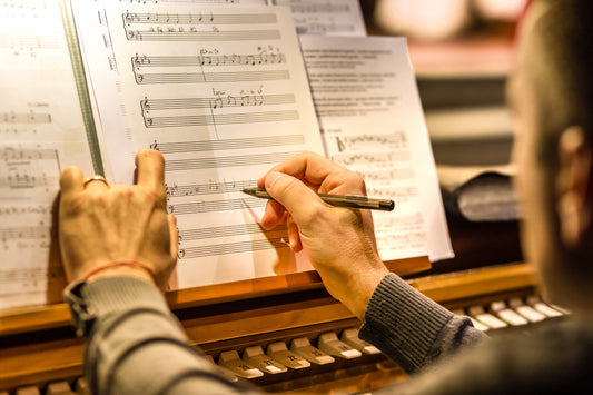 Organist's hands writing notes with a pen on sheet music resting on a warm wooden organ music rack above the pistons