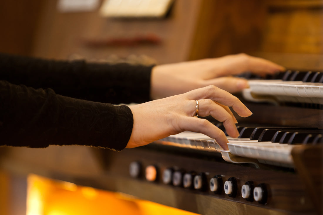 Organist's hands playing the manuals of a warm wooden church organ console, with piston buttons below the keys