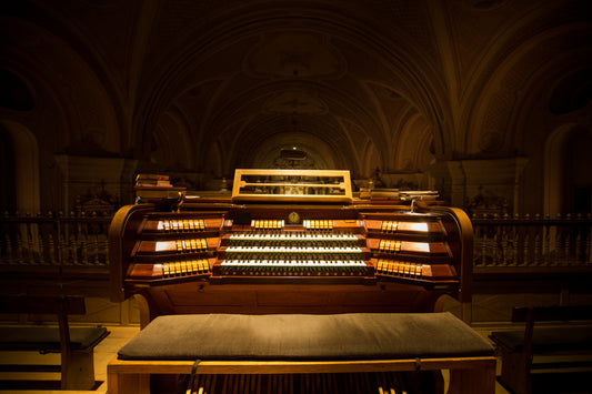 An empty multi-manual pipe organ console glowing in warm light in a dark, vaulted church, with the organ bench in the foreground