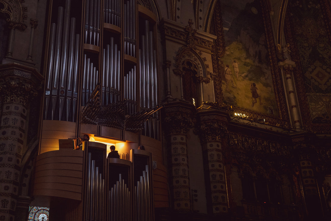 Organists seated at a softly lit gallery console in a dim cathedral, with tall silver organ pipes on the left and a painted mural on the right